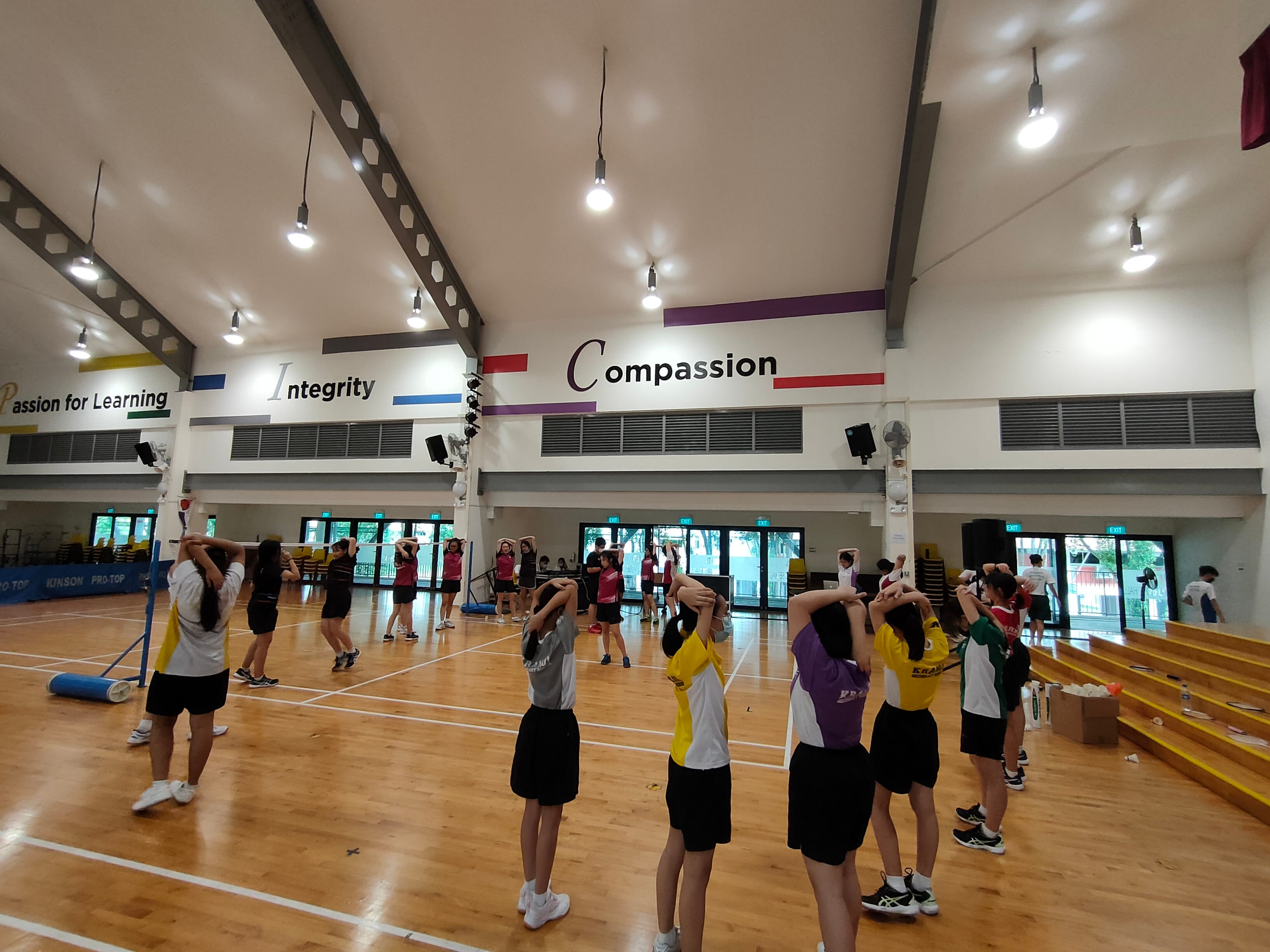 Students stretch with arms raised in a school gymnasium with words Integrity, Compassion, and Passion for Learning on the walls.